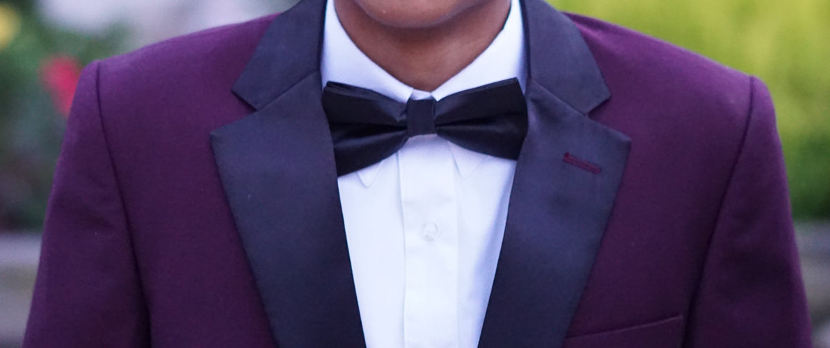 A close-up of a person wearing a formal tuxedo jacket in deep purple with black satin lapels, a white dress shirt, and a black bow tie.